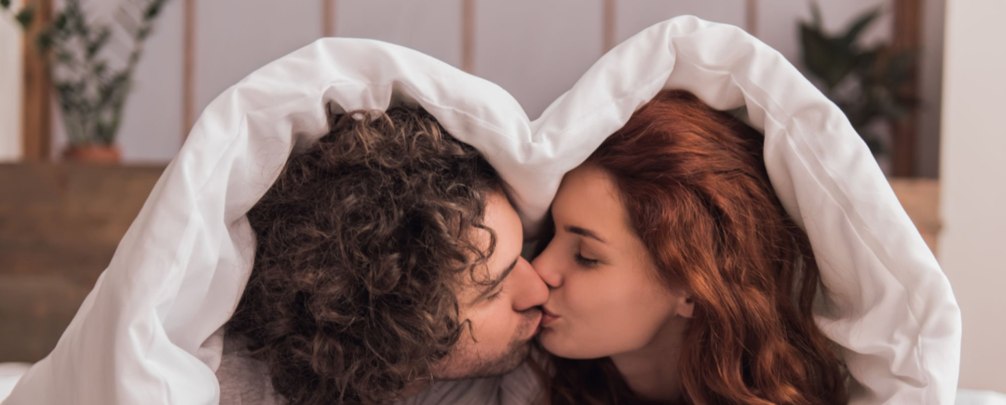 Couple kissing under a white blanket in a cozy indoor setting